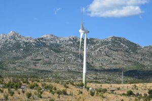 A lone wind turbine set against a backdrop of rugged mountains under a clear blue sky.