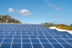 Expansive solar farm with rows of panels under clear blue sky and hills in the background.